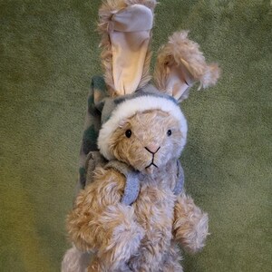 May include: A plush bunny wearing a blue and green hat and a grey backpack, standing on a pair of wooden snowshoes with red straps.