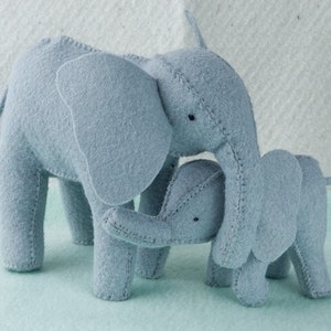 May include: Two gray felt elephant plushies, one larger than the other, are standing close together. The larger elephant has its trunk wrapped around the smaller elephant's neck.