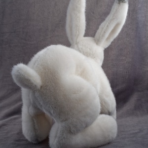 May include: A white plush bunny rabbit toy with gray ears. The bunny is sitting with its head down and its back to the camera.