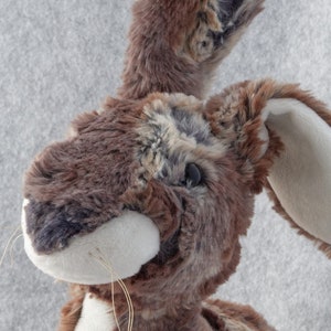 Könnte beinhalten: Ein Plüschtierhase mit detailliertem Fellmuster in Braun-, Grau- und Cremetönen. Der Hase hat lange Ohren, eine weiße Schnauze und zarte Schnurrhaare. Das Spielzeug ist mit realistischen Merkmalen und einer weichen, kuscheligen Textur gestaltet.