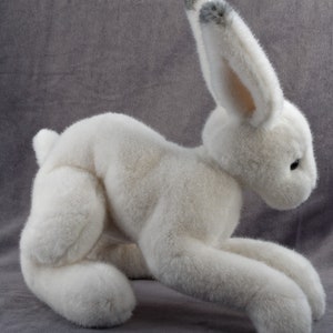 May include: A white plush rabbit toy with gray fur on its ears. The rabbit is lying on its belly with its front paws extended.