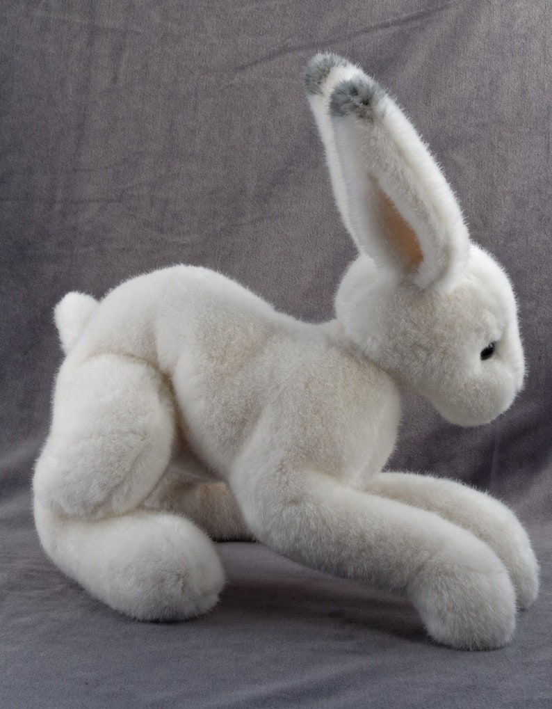 May include: A white plush rabbit toy with gray fur on its ears. The rabbit is lying on its belly with its front paws extended.