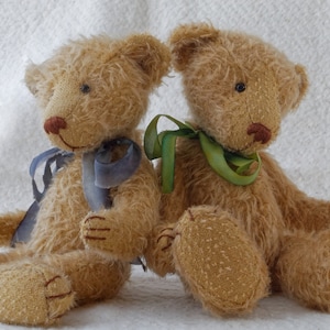 May include: Two plush teddy bears, one with a blue ribbon and the other with a green ribbon, sitting side by side on a white fluffy surface.