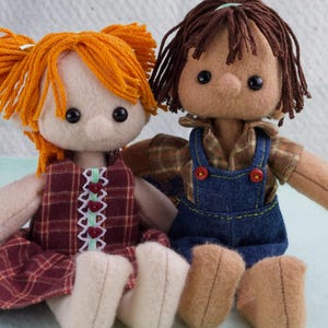 May include: Two handmade fabric dolls. One doll has orange yarn pigtails and a maroon plaid dress with white and green trim. The other doll has brown yarn hair and wears a brown plaid shirt with blue denim overalls. Both dolls have black button eyes.