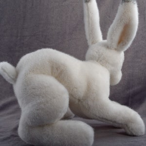 May include: A white plush rabbit toy with gray ears. The rabbit is sitting on a gray surface and is looking to the right.