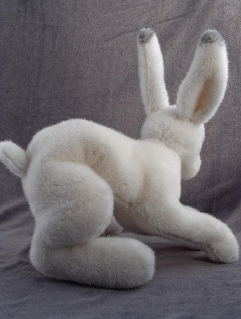May include: A white plush rabbit toy with gray ears. The rabbit is sitting on a gray surface and is looking to the right.