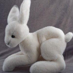 May include: A white plush rabbit toy with long ears and a fluffy tail. The rabbit is lying on its belly with its front paws extended and its back paws tucked under its body.