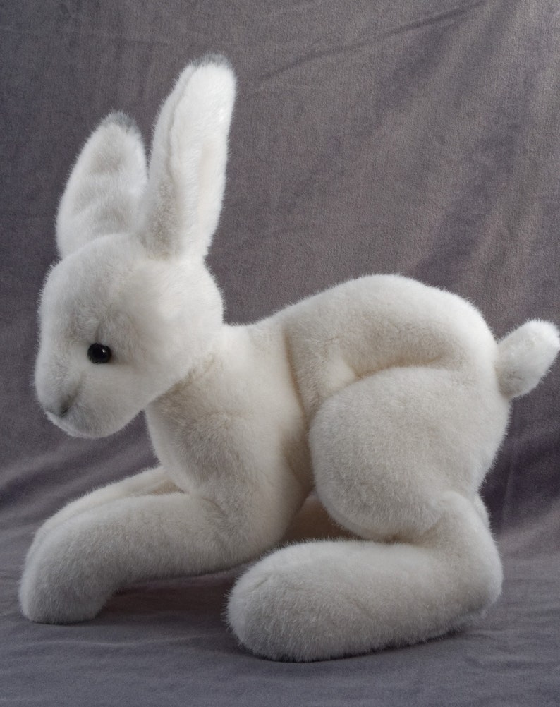 May include: A white plush rabbit toy with long ears and a fluffy tail. The rabbit is lying on its belly with its front paws extended and its back paws tucked under its body.