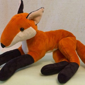May include: A plush fox toy featuring vibrant orange fur on its body, head, and tail. It has a white chest and tail tip, black ears, and black lower legs. The fox has a pointed snout with a small black nose and dark, round plastic eyes.