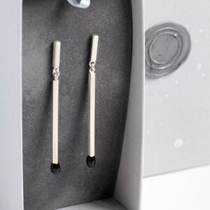 May include: A pair of silver earrings with black accents in a white box. The earrings are long and thin, with a simple design.