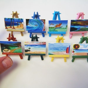 May include: A set of nine miniature easels with colorful wooden frames and painted canvas. The paintings depict various beach scenes with palm trees, waves, and sunsets. The easels are arranged in three rows of three.