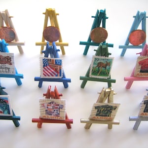 Op de afbeelding: Tien miniatuurhouten easels, geschilderd in verschillende kleuren, elk met een andere vintage postzegel. De postzegels tonen een verscheidenheid aan ontwerpen, waaronder een rood, wit en blauwe vlag, een hert en een Hawaiiaans shirt.