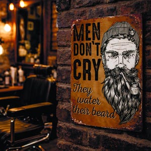 Könnte beinhalten: Ein Vintage-Metallschild mit dem Text "MEN DON'T CRY, They water their beard." Das Schild zeigt eine Schwarz-Weiß-Illustration eines bärtigen Mannes, der eine Pfeife raucht. Das Schild ist an einer Backsteinmauer befestigt, möglicherweise in einem Friseursalon.