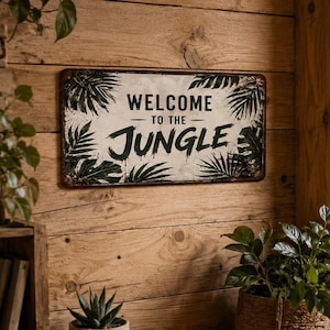 Puede incluir: Letrero metálico rectangular con acabado envejecido, con las palabras "WELCOME TO THE JUNGLE" en negro. El letrero está decorado con hojas de palma verdes y tiene un aspecto vintage y rústico.