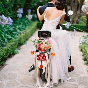 May include: A newly married couple rides a vintage scooter adorned with flowers and a "Just Married" sign. The bride wears a white wedding dress with lace. The scooter features a red taillight and orange indicators.