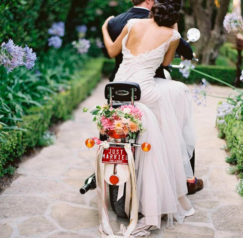 May include: A newly married couple rides a vintage scooter adorned with flowers and a "Just Married" sign. The bride wears a white wedding dress with lace. The scooter features a red taillight and orange indicators.