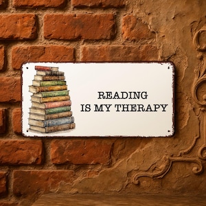 Könnte beinhalten: Rechteckiges Metallschild mit dem Text "READING IS MY THERAPY" in Schwarz. Das Schild zeigt einen Stapel Bücher mit farbigen Buchrücken. Das Schild ist an einer Backsteinmauer befestigt.