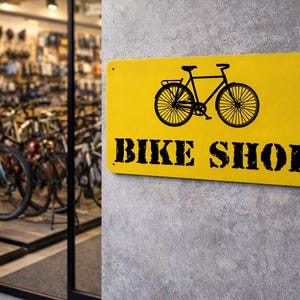 Könnte beinhalten: Ein gelbes rechteckiges Schild mit einer schwarzen Fahrradgrafik und dem Schriftzug "BIKE SHOP" in fetten, schwarzen Buchstaben. Das Schild ist an einer grauen Betonmauer befestigt, im Hintergrund ist ein Fahrradladen zu sehen.