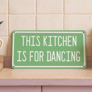 Może przedstawiać: Zielony metalowy znak z białym napisem "THIS KITCHEN IS FOR DANCING". Znak znajduje się na drewnianym blacie, z wazonem lawendy i białym dzbanem po obu stronach.