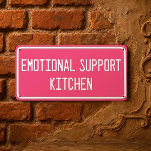 Op de afbeelding: Een roze rechthoekig bord met de woorden "EMOTIONAL SUPPORT KITCHEN" in witte letters. Het bord heeft een witte rand en is bevestigd aan een bakstenen muur. De bakstenen muur is een mix van rode en bruine bakstenen.