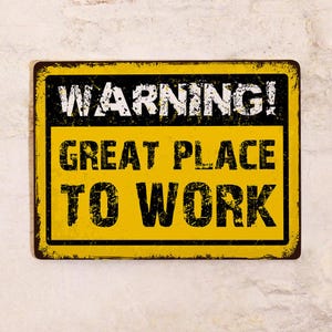 May include: A rectangular metal sign with a yellow background and black border. The sign has the words "WARNING! GREAT PLACE TO WORK" in black, distressed lettering. The sign has a weathered appearance.