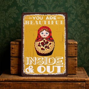 Pode incluir: Uma placa de metal retangular com um acabamento desgastado. A placa apresenta uma ilustração de uma boneca russa em vermelho, castanho e amarelo, com o texto "YOU ARE BEAUTIFUL INSIDE & OUT".