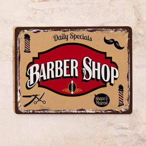 Może przedstawiać: Metalowy szyld w stylu vintage dla zakładu fryzjerskiego. Znak zawiera słowa "BARBER SHOP" pogrubioną białą czcionką na czerwono-czarnym tle. Zawiera słupy fryzjerskie, wąsy oraz tekst "Daily Specials" i "Shave & Haircut".