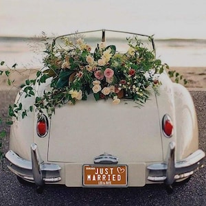 May include: A classic cream-coloured convertible adorned with a large floral arrangement of pink, white, and green flowers. The number plate reads "JUST MARRIED" with the names "LEO & KATE". The car is parked near the sea.