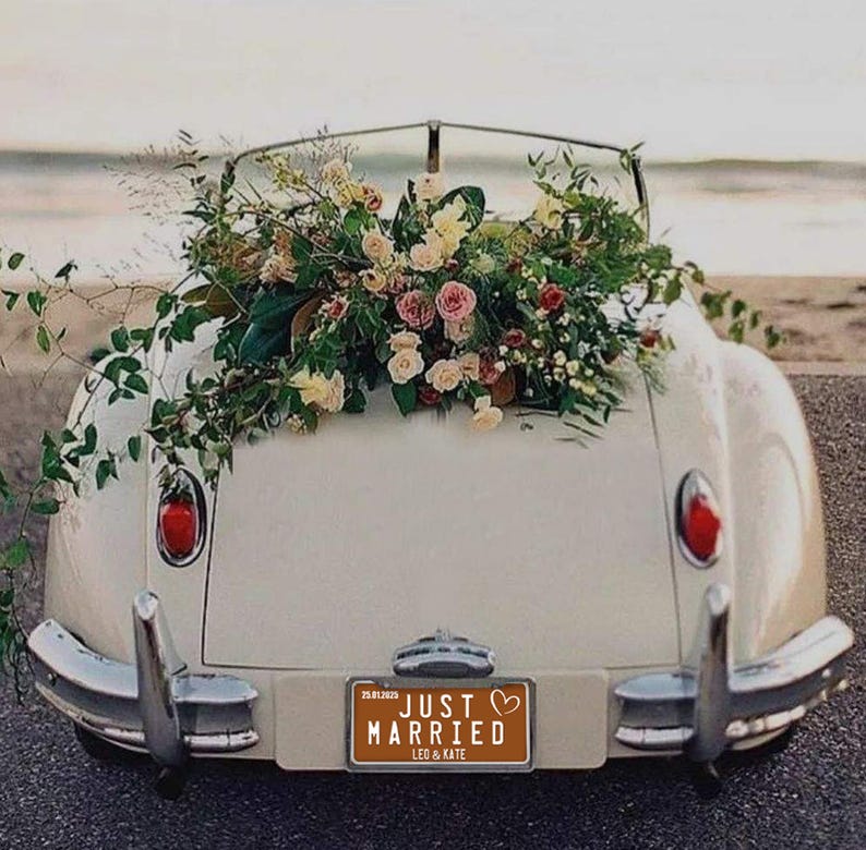 May include: A classic cream-coloured convertible adorned with a large floral arrangement of pink, white, and green flowers. The number plate reads "JUST MARRIED" with the names "LEO & KATE". The car is parked near the sea.