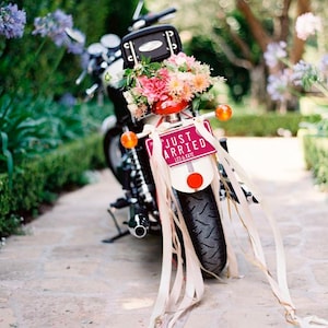 May include: A decorated motorcycle, ideal for a wedding. It has a bouquet of pink and peach flowers and a license plate that reads "JUST MARRIED" with the names "LEO & KATE". Cream ribbons are attached to the back.