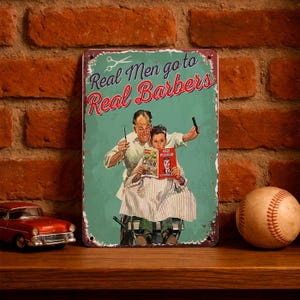 Puede incluir: Letrero metálico vintage con el texto "Real Men go to Real Barbers", que representa a un barbero y un cliente. El letrero tiene un aspecto envejecido con un fondo turquesa y texto rojo y blanco. También se ven una pelota de béisbol y un coche de juguete.