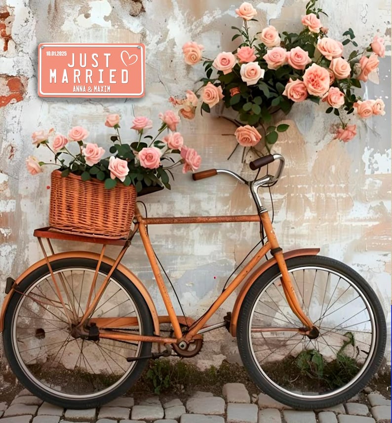 May include: An orange vintage bicycle with a wicker basket of pink roses. A sign reading "JUST MARRIED" is on the wall. The bicycle is positioned in front of a textured wall adorned with more pink roses.