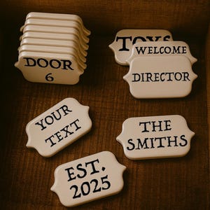 Puede incluir: Conjunto de etiquetas de cerámica blanquecina con texto negro. Las etiquetas muestran frases como "DOOR 6", "TOYS", "WELCOME", "DIRECTOR", "YOUR TEXT", "THE SMITHS" y "EST. 2025". Las etiquetas están dispuestas sobre una superficie de madera oscura.