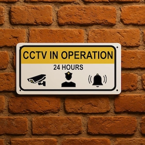 May include: A rectangular white sign with black text and icons, mounted on a brick wall. The sign reads "CCTV IN OPERATION 24 HOURS" with a camera, a police officer, and a bell icon.