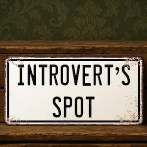 May include: A rectangular metal sign with the words "INTROVERT'S SPOT" in black, bold letters on a white background. The sign has a distressed, vintage look with a black border and rust-colored accents. It rests on a wooden surface.