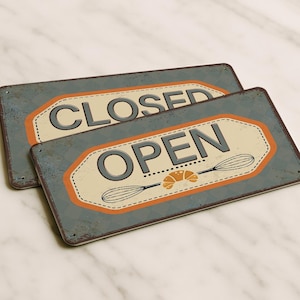 以下が含まれることがあります： 使い古した仕上げの長方形の金属看板が2つあります。1つの看板には「CLOSED」、もう1つには「OPEN」と書かれています。「OPEN」の看板には泡立て器とクロワッサンのグラフィックが描かれています。どちらの看板もオレンジ色の縁取りがあり、大理石の表面にあります。