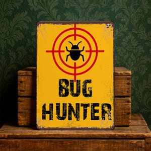 May include: A rectangular yellow metal sign with a distressed finish. The sign features a red target with a black insect silhouette at the centre. Below the target, the words "BUG HUNTER" are printed in black, distressed font.