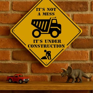 Könnte beinhalten: Ein gelbes, rautenförmiges Schild mit dem Text "IT'S NOT A MESS IT'S UNDER CONSTRUCTION" hängt an einer Backsteinmauer. Ein roter Spielzeug-LKW und eine graue Triceratops-Figur stehen auf einem Holzregal.