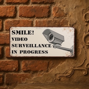 Pode incluir: Uma placa de metal retangular com o texto "SMILE! VIDEO SURVEILLANCE IN PROGRESS" e uma ilustração de uma câmera de vigilância cinza. A placa está montada em uma parede de tijolos com uma aparência vintage desgastada.