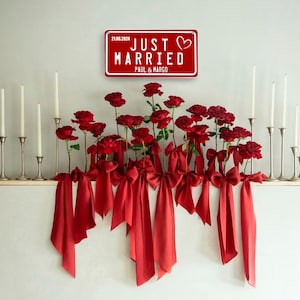 May include: A red sign reading "JUST MARRIED" with a heart symbol, the date 21.06.2024, and the names PAUL & MARGO. Below, red roses are adorned with red bows and ribbons. Silver candle holders with white candles are on either side.