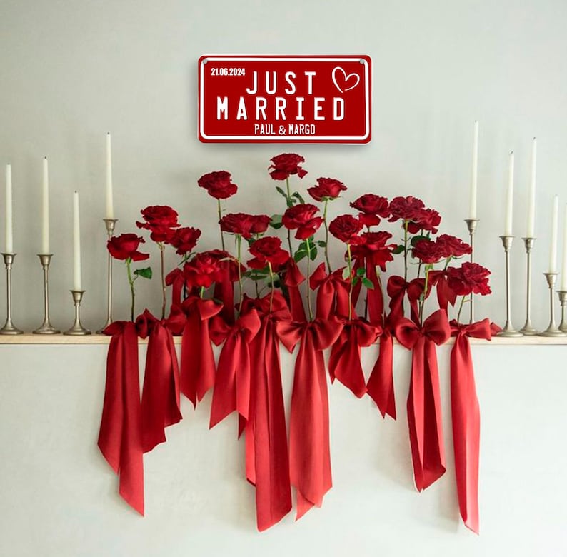 May include: A red sign reading "JUST MARRIED" with a heart symbol, the date 21.06.2024, and the names PAUL & MARGO. Below, red roses are adorned with red bows and ribbons. Silver candle holders with white candles are on either side.