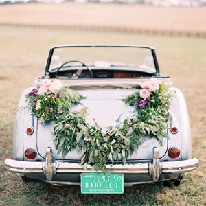 May include: A classic silver convertible adorned with flowers for a wedding. The car features a green number plate with the words "Just Married" and the names "Margarita & Roberto". The car is parked on grass.