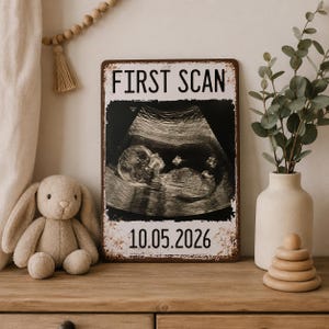May include: A distressed metal sign displaying "FIRST SCAN" and the date "10.05.2026", alongside a black and white ultrasound image. A soft toy bunny and a vase with foliage are visible in the background.
