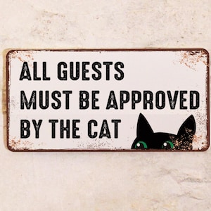 Könnte beinhalten: Rechteckiges Metallschild mit dem Text "ALL GUESTS MUST BE APPROVED BY THE CAT" in schwarzen Buchstaben. Eine schwarze Katze lugt aus der unteren rechten Ecke mit grünen Augen hervor. Das Schild hat einen verwitterten Vintage-Look mit rostfarbenen Rändern.