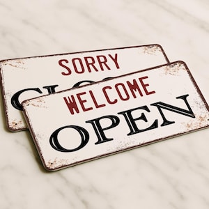 Peut inclure: Deux enseignes rectangulaires en métal aux bords usés. L'une porte l'inscription "SORRY" en lettres bordeaux. L'autre indique "WELCOME OPEN" en lettres bordeaux et noires. Les enseignes ont une esthétique vintage et rustique.
