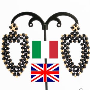 Puede incluir: Un par de pendientes ovalados negros y dorados con un diseño de cuentas. Los pendientes cuelgan de un soporte negro con una bandera italiana y británica detrás.
