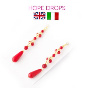 May include: A pair of long, red and gold earrings with a teardrop-shaped red stone at the bottom. The earrings are made with small, round beads and are strung on a gold wire.