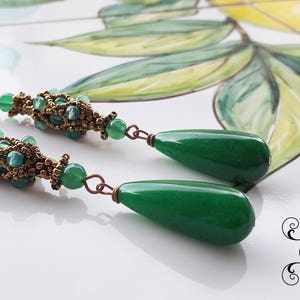 May include: A pair of green gemstone drop earrings with gold-toned bead accents. The earrings feature a teardrop-shaped gemstone and a beaded section with a woven design.