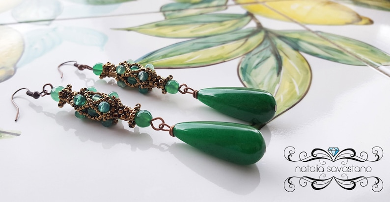 May include: A pair of green gemstone drop earrings with gold-toned bead accents. The earrings feature a teardrop-shaped gemstone and a beaded section with a woven design.