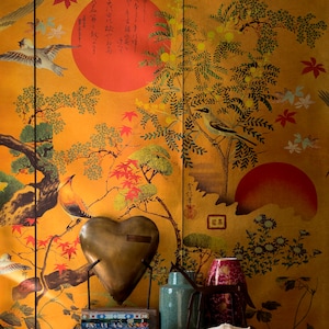 May include: A gold heart-shaped decorative object sits on a black table with a stack of books and a vase. The background is a gold wallpaper with a Japanese-inspired design featuring birds, trees, and flowers.